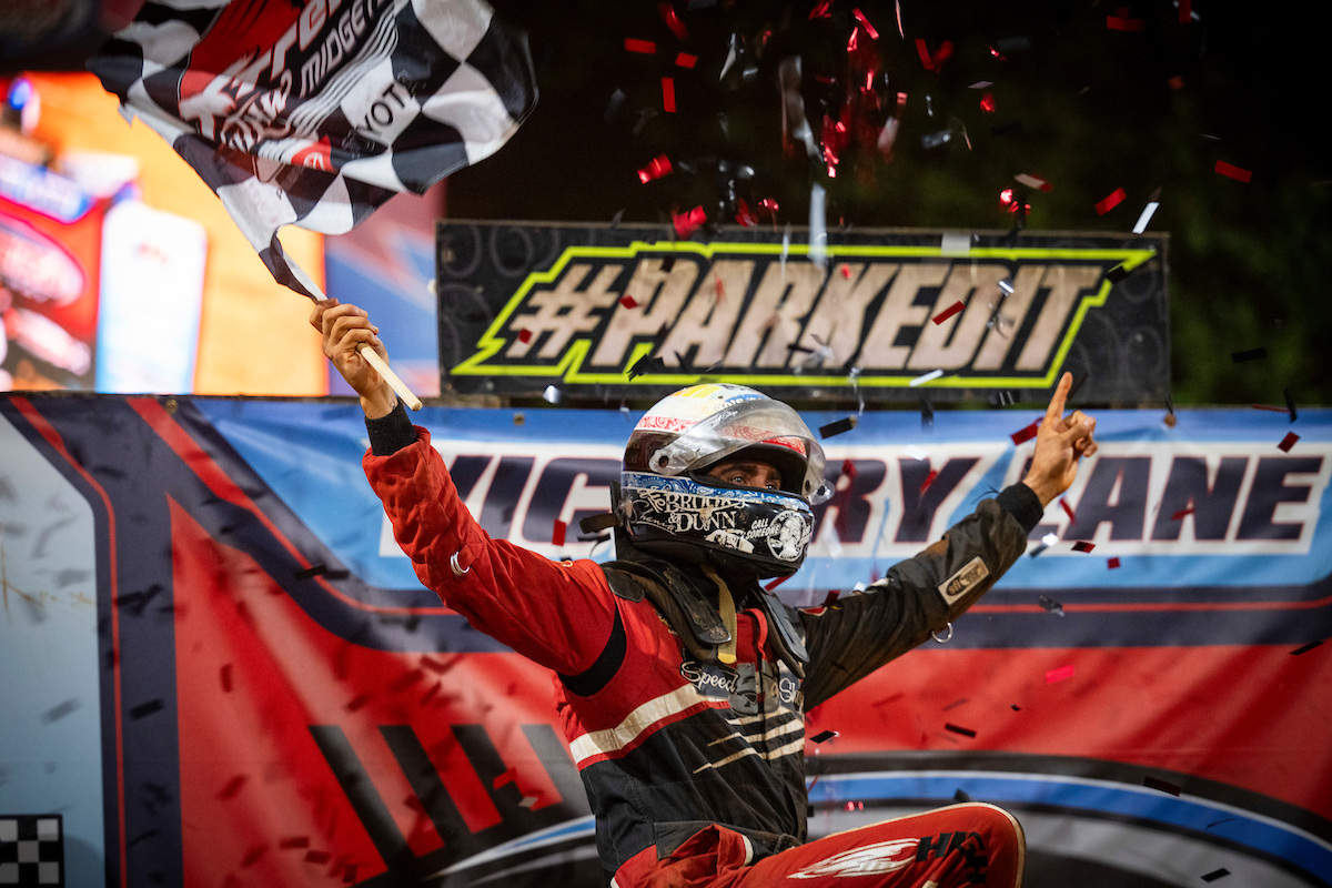 A DIRTcar Xtreme Outlaw Midget driver celebrates in victory lane with a checkered flag and confetti after winning a dirt racing event. 